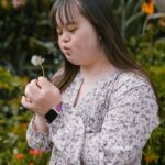 A young woman with Down syndrome holding and blowing a dandelion in a garden setting.